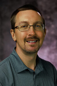 Man with glasses and a purple background smiling.