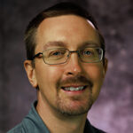 Man with glasses and a purple background smiling.